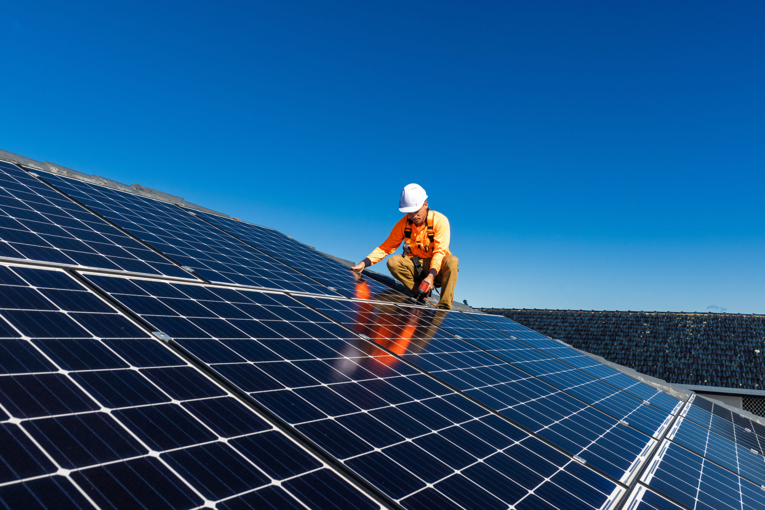 Solar panel technician with drill installing solar panels on house roof on a sunny day.